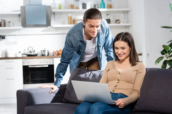 Jeune homme debout près de copine excitée assis sur le canapé dans la cuisine et à l'aide d'un ordinateur portable — Photo de stock