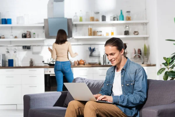 Foco seletivo de homem animado usando laptop no sofá na cozinha e namorada de pé no fundo — Fotografia de Stock