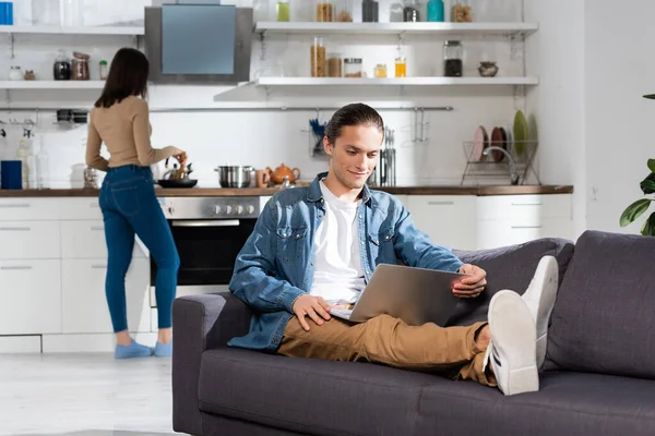 Foco seletivo do homem usando laptop no sofá na cozinha e mulher de pé no fundo — Fotografia de Stock