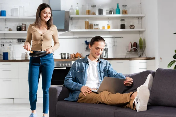 Joven usando el ordenador portátil en el sofá en la cocina, mientras que la novia de pie con la pluma de freír y espátula — Stock Photo