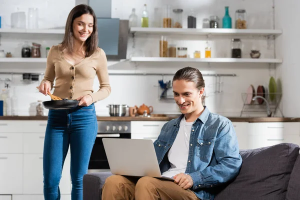 Brunette woman holding frying pen and spatula while standing near boyfriend using laptop on sofa — Stock Photo