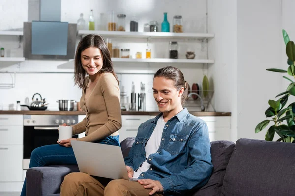 Brünette Frau mit Tasse Kaffee sitzt auf Sofa in Küche in der Nähe Freund mit Laptop — Stockfoto