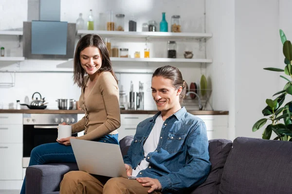 Morena mujer con taza de café sentado en el sofá en la cocina cerca de novio utilizando el ordenador portátil - foto de stock