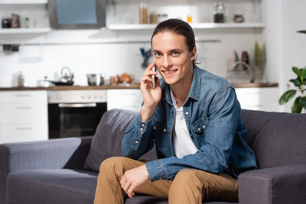 Молодий чоловік в джинсовій сорочці дивиться на камеру, сидячи на кухні і розмовляючи на смартфоні — стокове фото