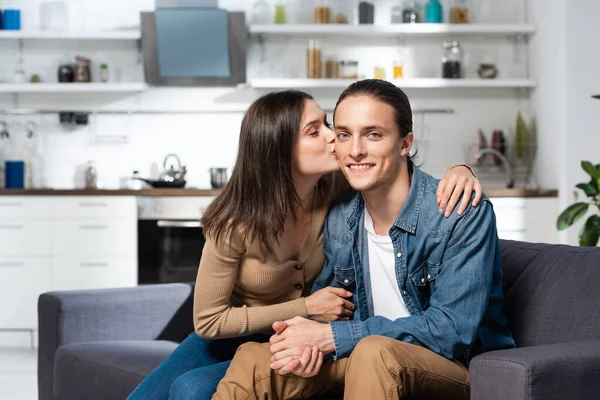 Brunette femme baisers joyeux homme assis sur canapé dans la cuisine et regardant caméra — Photo de stock