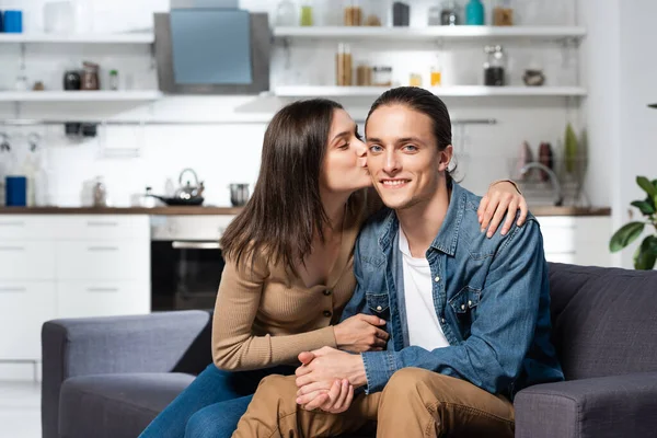 Brünette Frau küsst freudigen Mann sitzt auf Sofa in Küche und schaut in die Kamera — Stockfoto