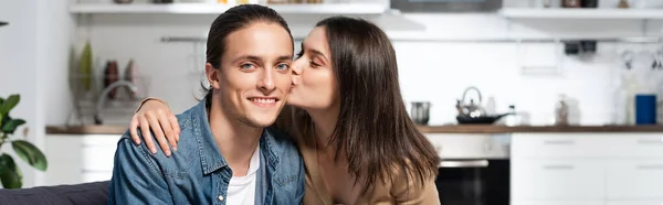 Orientación panorámica de la mujer morena besando novio complacido mirando a la cámara en la cocina - foto de stock