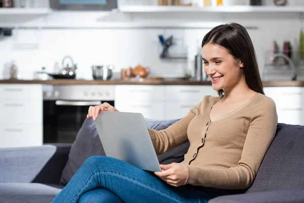 Freelance gioioso utilizzando il computer portatile mentre seduto sul divano in cucina — Foto stock