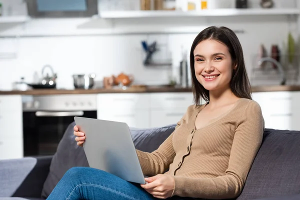 Emocionado freelancer mirando a la cámara mientras está sentado en el sofá en la cocina con el ordenador portátil — Stock Photo