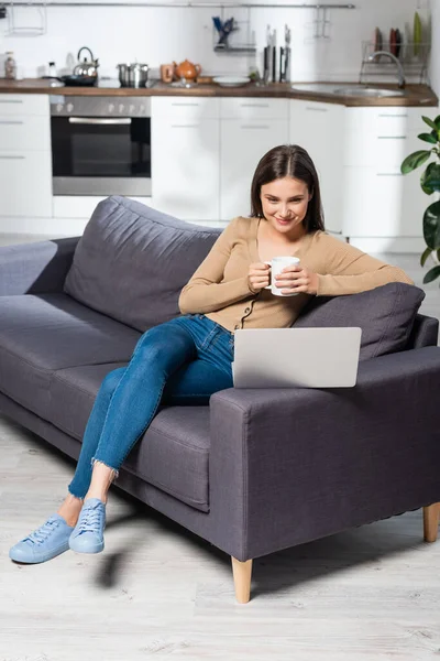 Задоволений фрілансер тримає чашку теплого напою, сидячи на дивані на кухні біля ноутбука — стокове фото