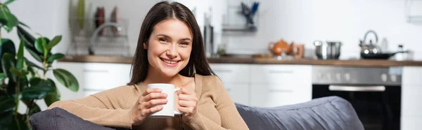 Concetto orizzontale di donna soddisfatta guardando la fotocamera mentre seduto sul divano in cucina con tazza di bevanda riscaldante — Foto stock