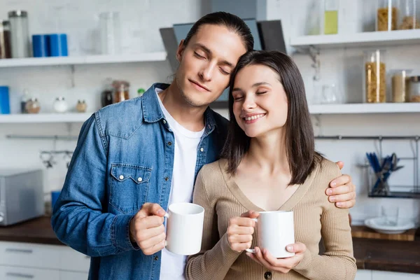 Pleased couple standing in kitchen with closed eyes while holding cups of coffee — Stock Photo