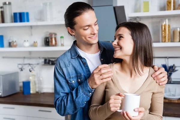 Молодий чоловік торкається плечей радісної дівчини, тримає чашку кави на кухні — стокове фото