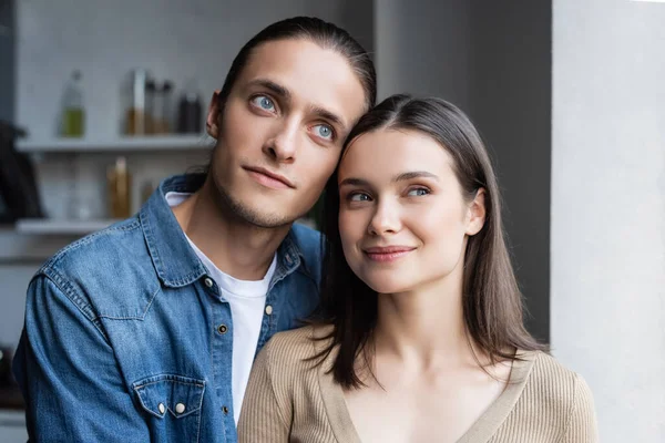 Pareja de ensueño mirando hacia otro lado mientras están de pie juntos en la cocina - foto de stock