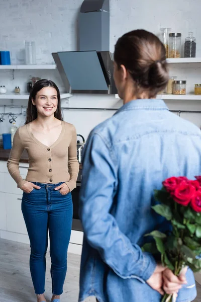 Vista trasera del joven sosteniendo rosas cerca de la mujer emocionada de pie con las manos en los bolsillos en la cocina - foto de stock