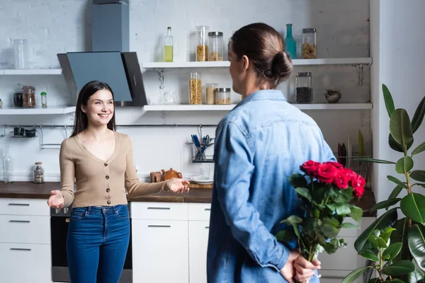 Rückansicht eines Mannes mit einem Rosenstrauß auf dem Rücken neben einer aufgeregten Frau, die mit offenen Armen in der Küche steht — Stockfoto