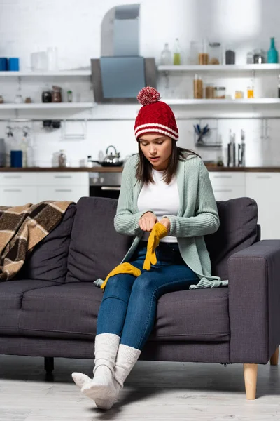 Femme gelée en tricot chapeau et chaussettes mettre des gants chauds tout en étant assis sur le canapé dans la cuisine froide — Photo de stock