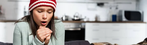 Horizontal image of freezing woman in knitted hat blowing on clenched hands in cold kitchen — Stock Photo