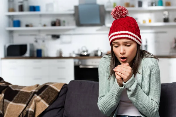 Cold woman in warm hat blowing on clenched hands while sitting on sofa in kitchen — Stock Photo