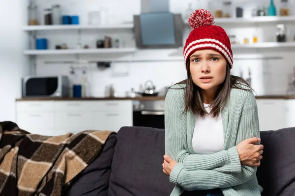 Freezing woman in knitted hat hugging herself while sitting in cold kitchen — Stock Photo