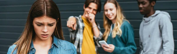 Panoramic shot of sad girl near laughing multiethnic teenagers with smartphone outdoors — Stock Photo