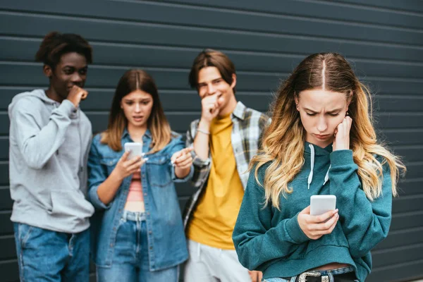 Foco seletivo de menina chateada usando smartphone perto de rir adolescentes multiétnicos ao ar livre — Fotografia de Stock