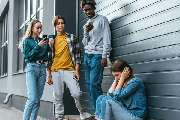 Selective focus of sad girl covering ears near multiethnic teenagers with smartphones and building — Stock Photo