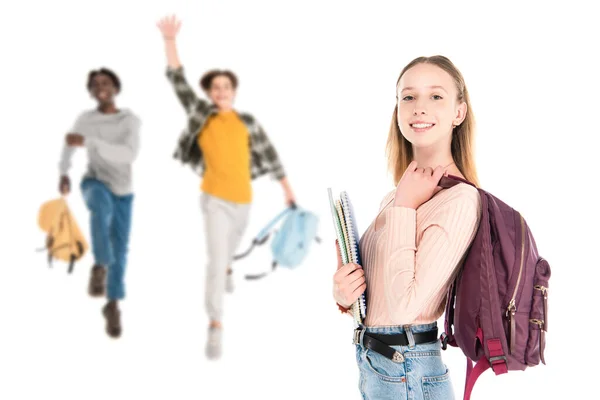Foco seletivo de adolescente sorridente segurando cadernos e mochila perto de amigos multiétnicos isolados em branco — Fotografia de Stock