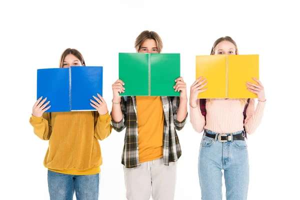 Teenagers covering faces with copy books and looking at camera isolated on white — Stock Photo