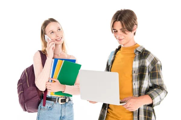 Улыбающийся подросток держит ноутбук рядом с другом с ноутбуками разговаривая на смартфоне изолированы на белом — стоковое фото