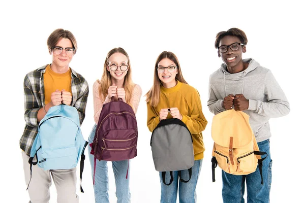 Smiling multicultural teenagers holding backpacks and looking at camera isolated on white — Stock Photo