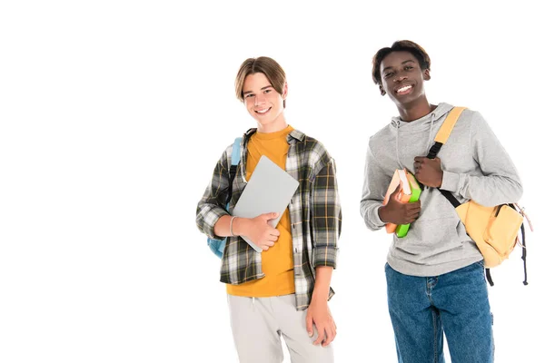 Lächelnde multiethnische Teenager mit Laptop und Büchern, die isoliert auf weiß in die Kamera schauen — Stockfoto