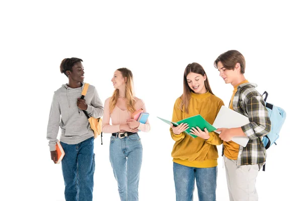 Sonrientes adolescentes multiétnicos con portátil y cuadernos hablando aislados en blanco - foto de stock