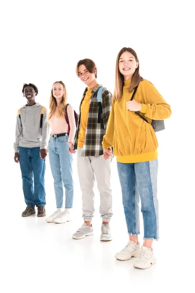 Adolescents multiethniques positifs avec des sacs à dos tenant la main et regardant la caméra sur fond blanc — Photo de stock