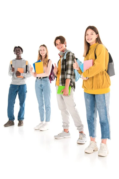 Adolescents multiculturels avec des livres et des sacs à dos souriant à la caméra et montrant comme sur fond blanc — Photo de stock