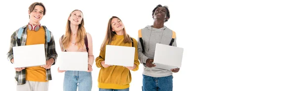 Cultivo panorámico de adolescentes multiculturales positivos que sostienen computadoras portátiles aisladas en blanco - foto de stock