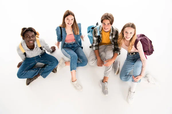 Visão aérea de adolescentes multiétnicos com mochilas sorrindo para a câmera no fundo branco — Fotografia de Stock