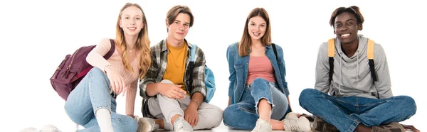 Foto panorámica de adolescentes positivos con mochilas sentadas sobre fondo blanco - foto de stock