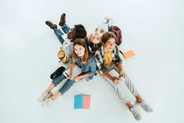Vista aérea de adolescentes multiétnicos sonriendo a la cámara mientras escriben en cuadernos sobre fondo gris - foto de stock