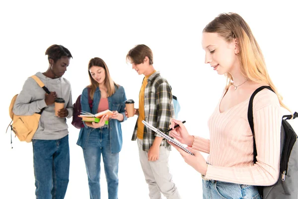 Enfoque selectivo de la escritura adolescente en el cuaderno cerca de amigos multiétnicos con libros y café para ir aislado en blanco - foto de stock