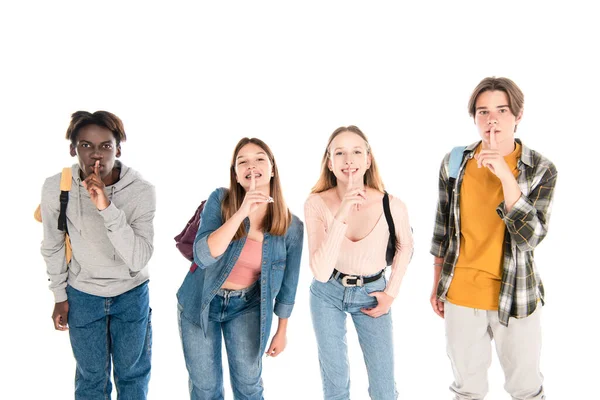 Sonrientes adolescentes multiétnicos con mochilas mostrando gesto shh aislado en blanco - foto de stock