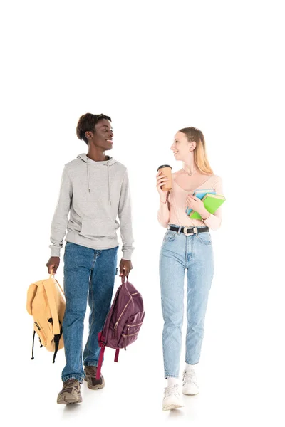 Улыбающийся африканский американский подросток держит рюкзаки рядом с другом с кофе, чтобы пойти и книги на белом фоне — стоковое фото