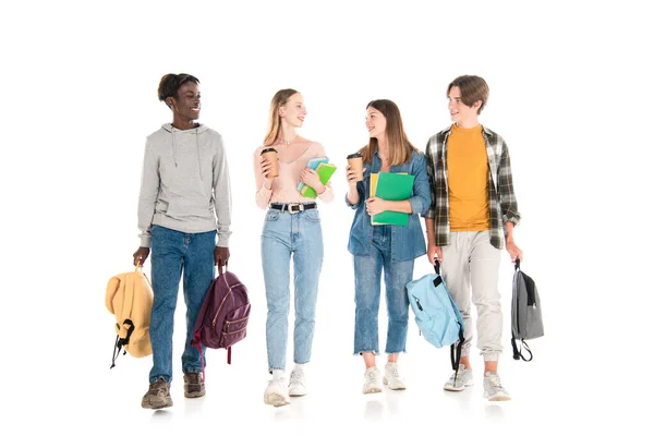 Adolescentes sonrientes multiculturales con café para llevar, libros y mochilas caminando sobre fondo blanco - foto de stock