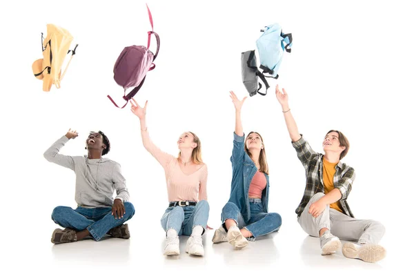 Позитивные мультиэтнические подростки бросают рюкзаки, сидя на белом фоне — стоковое фото