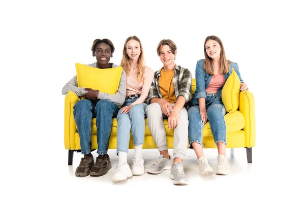 Multiethnische Teenager lächeln in die Kamera, während sie auf der Couch auf weißem Hintergrund sitzen — Stockfoto