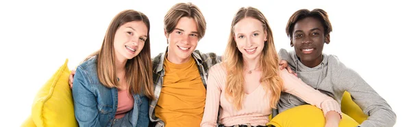 Panoramic crop of smiling multicultural teenagers looking at camera on yellow sofa isolated on white — Stock Photo