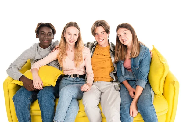 Позитивные мультиэтнические подростки смотрят в камеру, сидя на желтом диване, изолированном на белом — стоковое фото