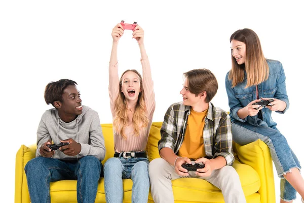 KYIV, UCRANIA - 21 de agosto de 2020: Adolescentes alegres y multiculturales jugando videojuegos en un sofá amarillo aislado en blanco - foto de stock