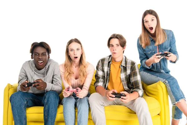 KYIV, UKRAINE - AUGUST 21, 2020: Excited multicultural friends holding joysticks on yellow couch isolated on white — Stock Photo