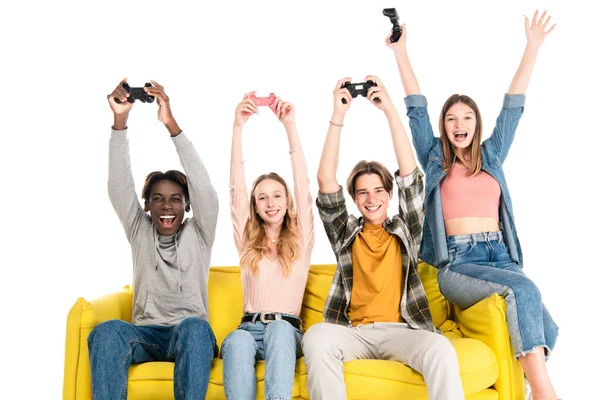 KYIV, UCRANIA - 21 de agosto de 2020: Adolescentes multiétnicos alegres sosteniendo joysticks en un sofá amarillo aislado en blanco - foto de stock